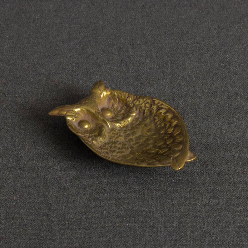 May include: A small, shallow brass dish shaped like an owl. The metal object features a textured surface that mimics feathers, with two prominent circular eyes and pointed ear tufts. The finish is a metallic gold tone with dark patina accents.