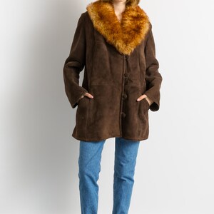 May include: A brown suede coat with a faux fur collar. The coat has a button-up front and pockets. The person is wearing blue jeans and burgundy ankle boots. The coat is a vintage style.