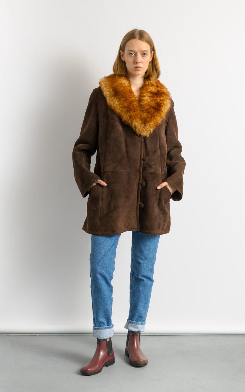 May include: A brown suede coat with a faux fur collar. The coat has a button-up front and pockets. The person is wearing blue jeans and burgundy ankle boots. The coat is a vintage style.
