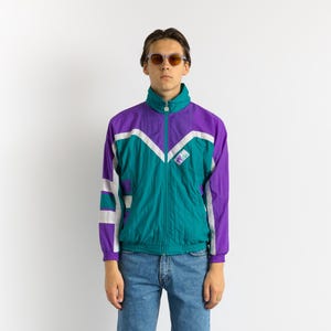 Könnte beinhalten: Eine Vintage-Trainingsjacke in Türkis, Lila und Weiß mit hohem Kragen und durchgehendem Reißverschluss. Die Jacke hat ein geometrisches Design auf der Vorderseite und den Ärmeln. Die Person im Bild trägt eine blaue Jeans und eine Sonnenbrille.