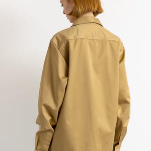 May include: A long-sleeved, tan button-down shirt with a relaxed fit. The shirt has a collar and a single button closure at the top.