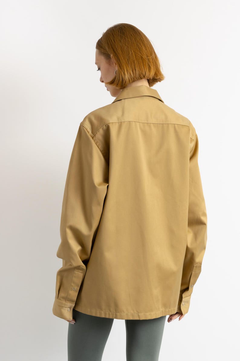 May include: A long-sleeved, tan button-down shirt with a relaxed fit. The shirt has a collar and a single button closure at the top.
