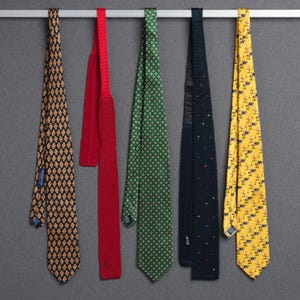 May include: Five neckties of various patterns and colors hang from a silver bar. The ties include a black tie with a repeating orange design, a solid red tie, a green tie with a white dot pattern, a navy tie with small colored dots, and a yellow tie with a repeating windmill pattern.