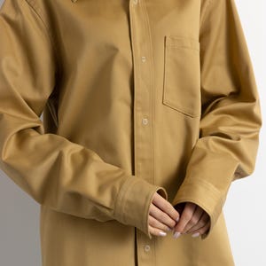 May include: A long-sleeved, oversized, light brown shirt with a button-down front and a single chest pocket.