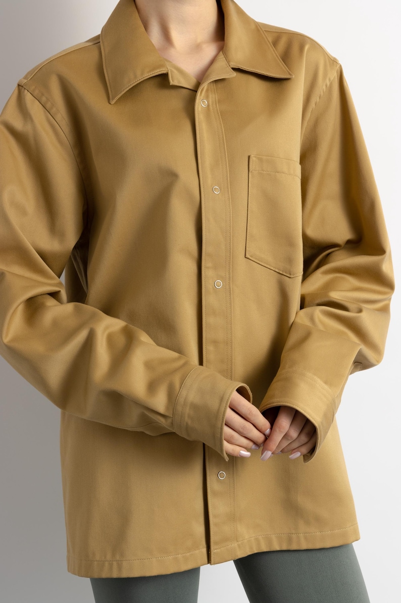 May include: A long-sleeved, oversized, light brown shirt with a button-down front and a single chest pocket.