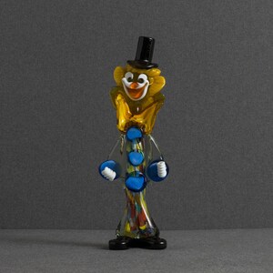 May include: A glass figurine of a clown wearing a black top hat, a yellow ruffled collar, and a long, slender body with blue circular accents. The clown has a white face with a painted orange nose and mouth, and black shoes at the base.