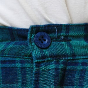 May include: Close-up of a blue button with the word "DOUGLAS" written on it. The button is sewn onto a pair of green and blue plaid pants.