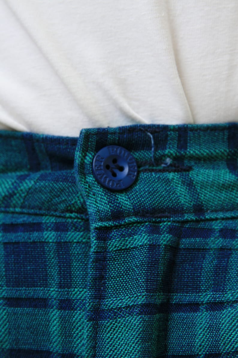 May include: Close-up of a blue button with the word "DOUGLAS" written on it. The button is sewn onto a pair of green and blue plaid pants.