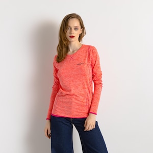 Puede incluir: Una camiseta deportiva de manga larga de color coral con la marca "CRAFT" en negro en el pecho. La camiseta tiene cuello redondo y ribete rosa en los puños y el dobladillo. La modelo lleva vaqueros azul oscuro.