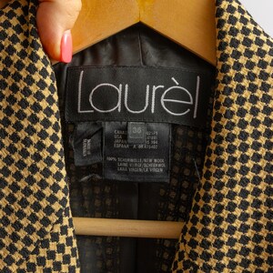 May include: Close-up of a black and gold patterned jacket with the brand name "Laur&egrave;l" visible. The jacket is hanging on a wooden hanger. The label indicates the jacket is made of 100% new wool.