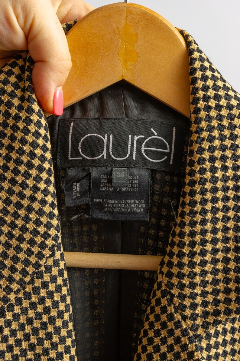 May include: Close-up of a black and gold patterned jacket with the brand name "Laur&egrave;l" visible. The jacket is hanging on a wooden hanger. The label indicates the jacket is made of 100% new wool.