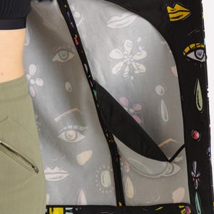 May include: A black jacket with a colorful, abstract print featuring eyes, lips, and flowers. The jacket has a mesh lining and a zipper closure.