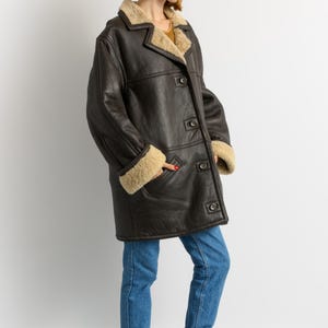 May include: A dark brown leather coat with a shearling lining and trim. The coat has a button-up front, pockets, and a classic collar. The sleeves have shearling cuffs. The person is wearing blue jeans and burgundy boots.