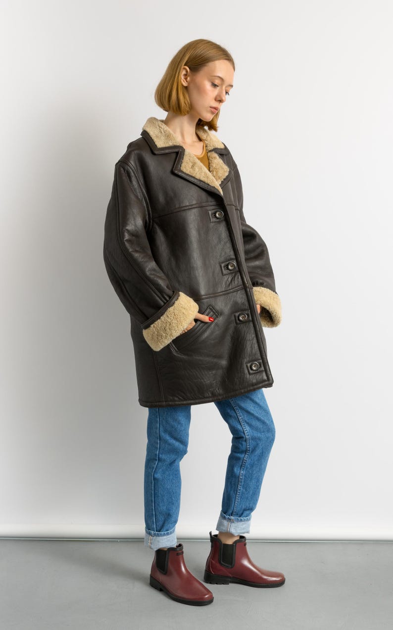 May include: A dark brown leather coat with a shearling lining and trim. The coat has a button-up front, pockets, and a classic collar. The sleeves have shearling cuffs. The person is wearing blue jeans and burgundy boots.