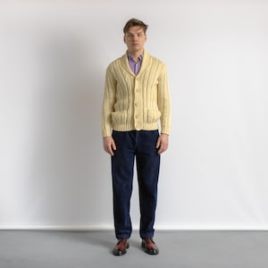 May include: A light yellow cable-knit cardigan with a shawl collar and button closure. The cardigan is worn over a purple collared shirt and paired with navy blue corduroy pants and brown leather shoes. The outfit is a classic, preppy style.