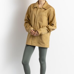 May include: A woman is wearing a khaki long-sleeved shirt with a button-up front and a chest pocket. She is also wearing gray leggings and red leather sandals with wooden soles.