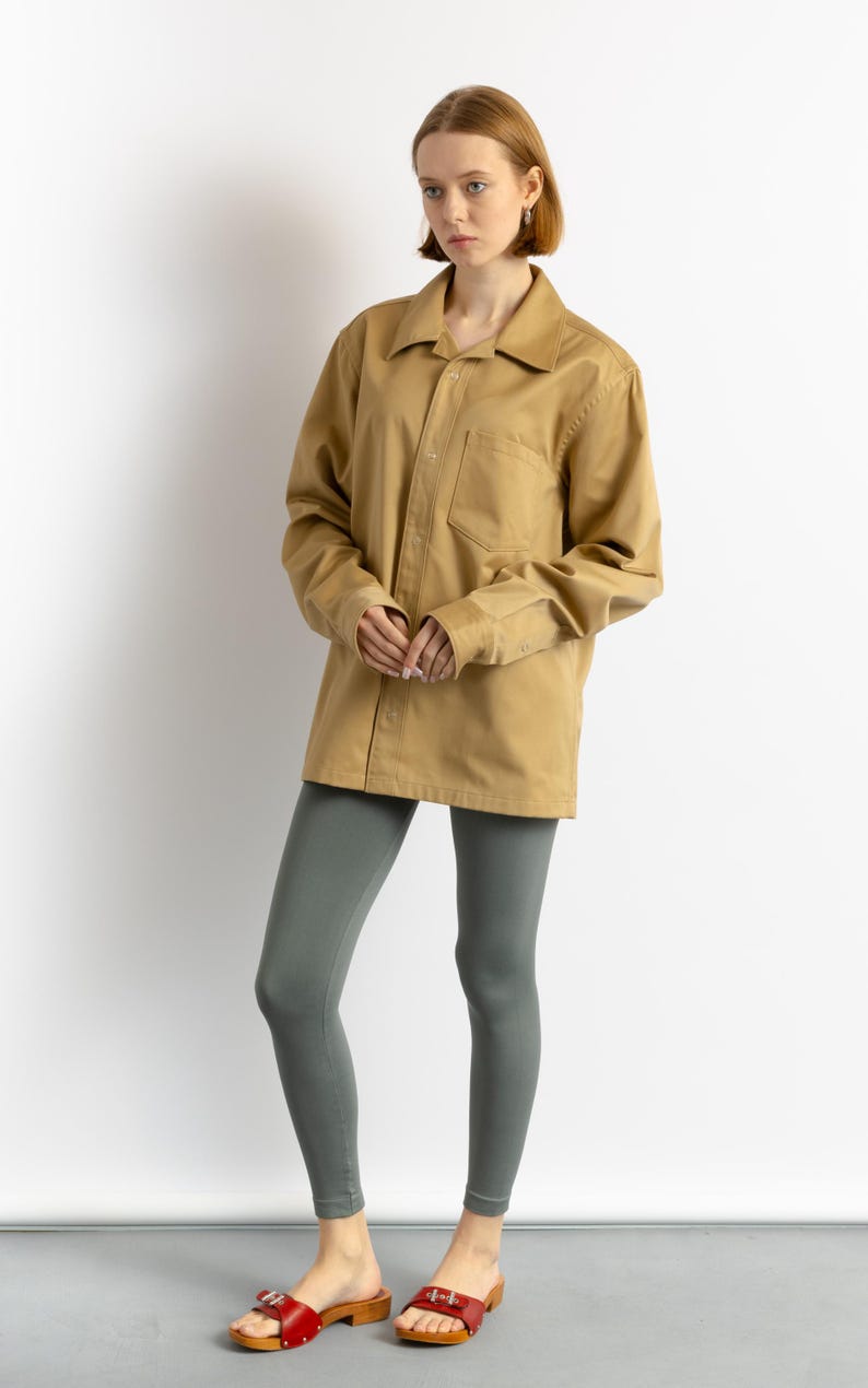May include: A woman is wearing a khaki long-sleeved shirt with a button-up front and a chest pocket. She is also wearing gray leggings and red leather sandals with wooden soles.
