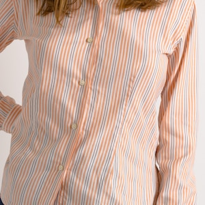 Puede incluir: Una camisa de manga larga con botones, con finas rayas naranjas y blancas. La camisa tiene un cuello cl&aacute;sico y una silueta ajustada.
