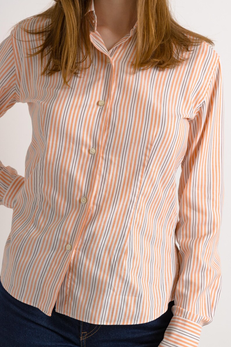 Puede incluir: Una camisa de manga larga con botones, con finas rayas naranjas y blancas. La camisa tiene un cuello cl&aacute;sico y una silueta ajustada.