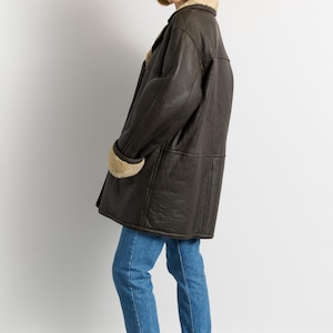 May include: A dark brown leather coat with a shearling collar and cuffs. The coat is knee-length and worn with blue jeans and burgundy rain boots. The coat has a classic, vintage style.