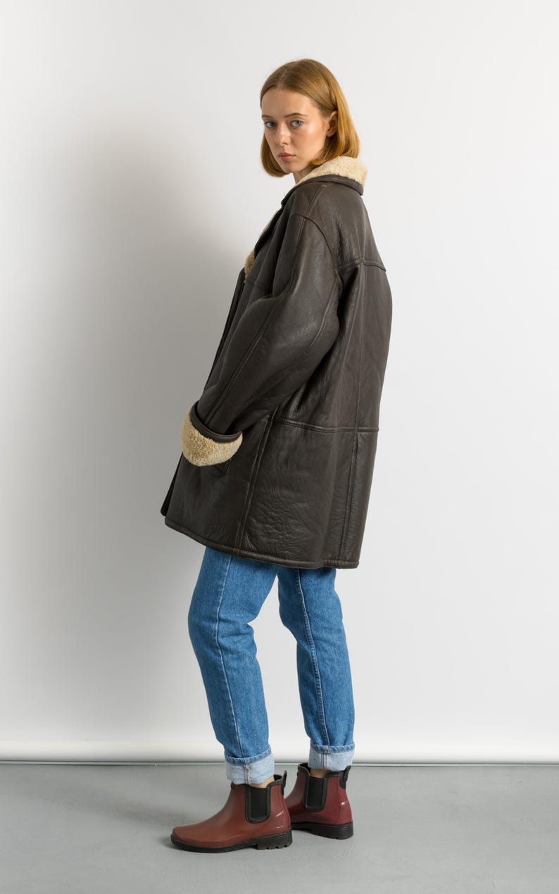 May include: A dark brown leather coat with a shearling collar and cuffs. The coat is knee-length and worn with blue jeans and burgundy rain boots. The coat has a classic, vintage style.