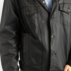 May include: A black leather jacket with a button-up front and a classic denim jacket style. The jacket has two flap pockets on the chest and two side pockets.
