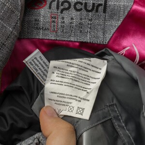 Puede incluir: Primer plano de la etiqueta de una chaqueta gris Rip Curl con informaci&oacute;n de tallas. La chaqueta tiene un forro de sat&eacute;n rosa y una etiqueta con detalles de composici&oacute;n de la tela. La etiqueta garantiza la autenticidad del producto.