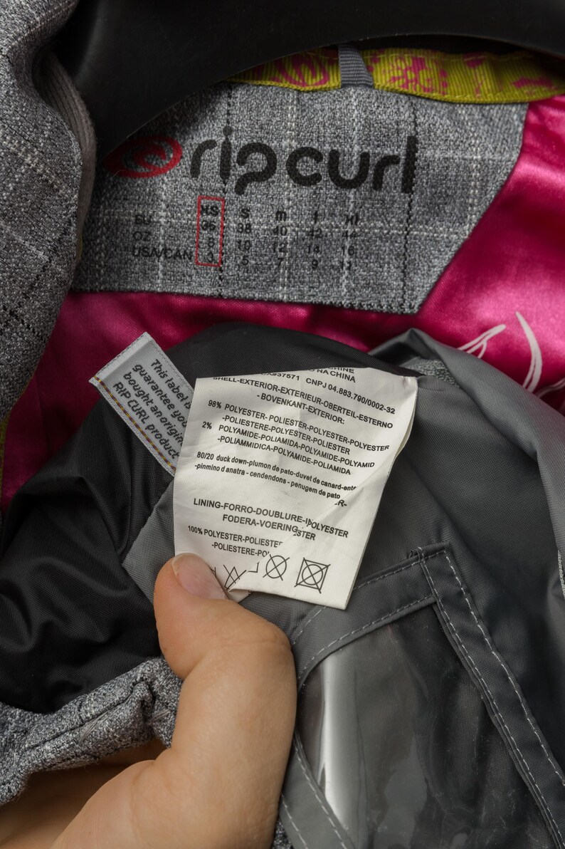 Puede incluir: Primer plano de la etiqueta de una chaqueta gris Rip Curl con informaci&oacute;n de tallas. La chaqueta tiene un forro de sat&eacute;n rosa y una etiqueta con detalles de composici&oacute;n de la tela. La etiqueta garantiza la autenticidad del producto.