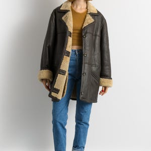 May include: A dark brown shearling coat with a tan interior and trim. The coat has a button-up front and a wide collar. The person in the image is wearing blue jeans and maroon boots.