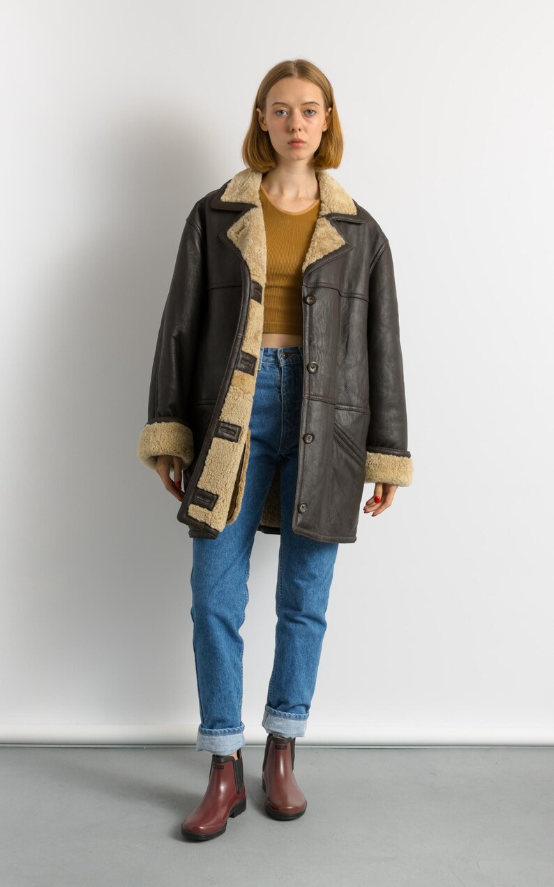 May include: A dark brown shearling coat with a tan interior and trim. The coat has a button-up front and a wide collar. The person in the image is wearing blue jeans and maroon boots.