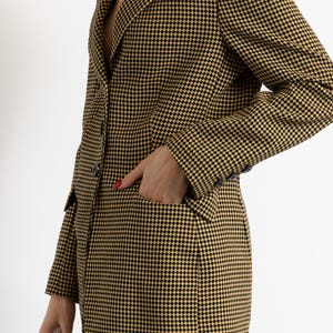 May include: A houndstooth blazer in black and gold. The blazer has a classic notched lapel, a two-button closure, and a pocket on the front. The sleeves have buttons at the cuffs. The blazer is worn over a top and blue jeans.