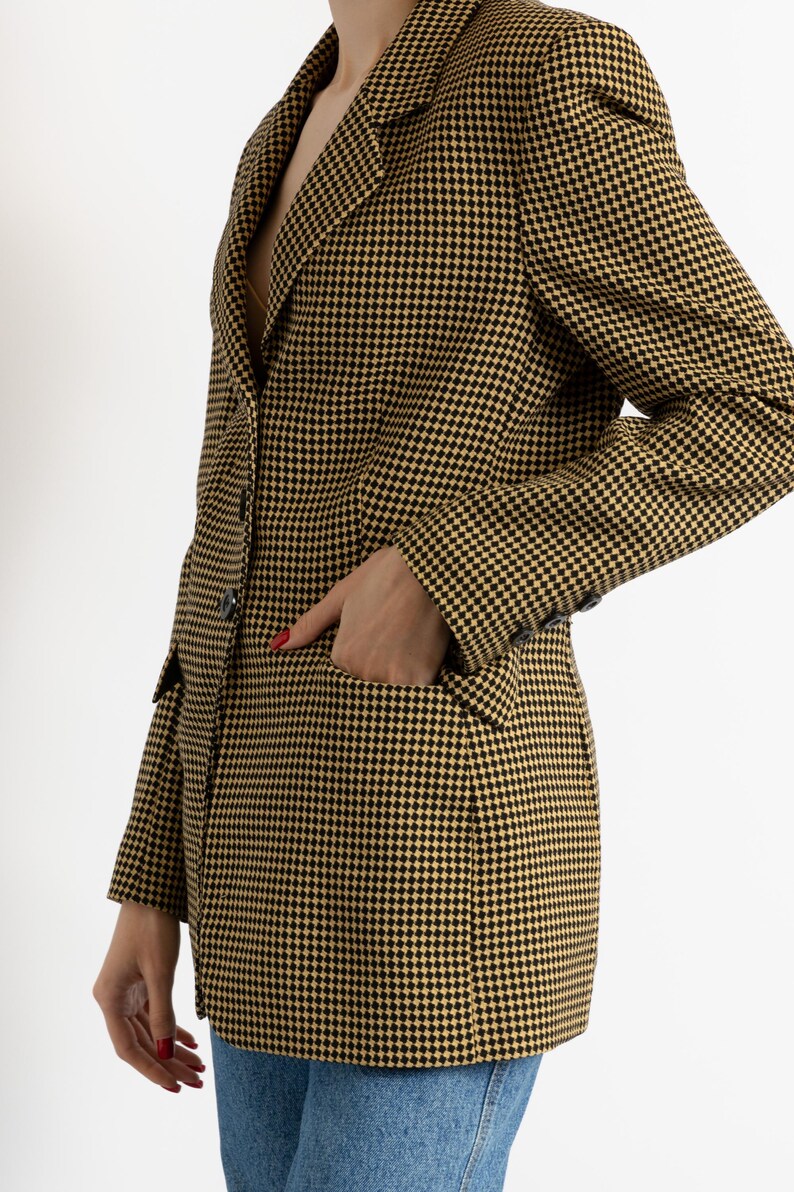 May include: A houndstooth blazer in black and gold. The blazer has a classic notched lapel, a two-button closure, and a pocket on the front. The sleeves have buttons at the cuffs. The blazer is worn over a top and blue jeans.