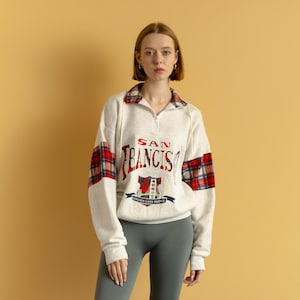 May include: A white sweatshirt with a red and black plaid pattern on the sleeves. The sweatshirt has a zipper closure and a "SAN FRANCISCO" graphic on the front.