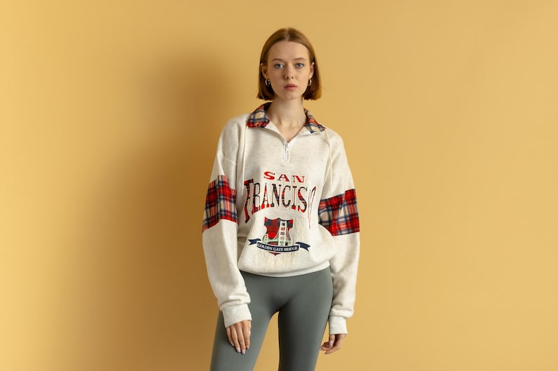 May include: A white sweatshirt with a red and black plaid pattern on the sleeves. The sweatshirt has a zipper closure and a "SAN FRANCISCO" graphic on the front.