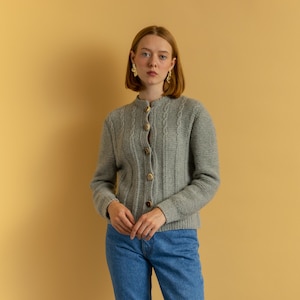 May include: A light gray cable-knit cardigan with a round neckline and long sleeves. The cardigan features a button-down front with five gold-colored buttons. The garment is made of a soft, textured material and is worn with blue jeans.