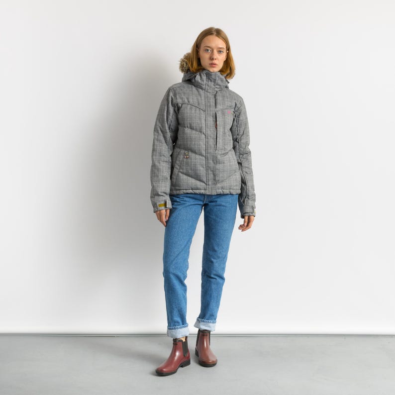 Puede incluir: Una chaqueta de invierno a cuadros gris y blanco con capucha forrada de piel. La chaqueta tiene cierre de cremallera, m&uacute;ltiples bolsillos y se usa con vaqueros azules y botas burdeos.