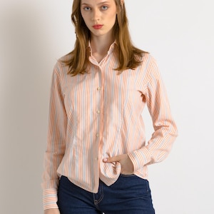 Puede incluir: Una mujer con una camisa de manga larga a rayas de color melocot&oacute;n y azul con fondo blanco. Lleva pantalones vaqueros azul oscuro y tiene la mano en el bolsillo.