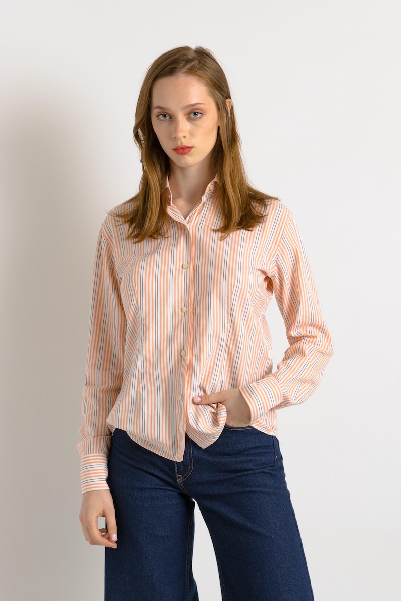 Puede incluir: Una mujer con una camisa de manga larga a rayas de color melocot&oacute;n y azul con fondo blanco. Lleva pantalones vaqueros azul oscuro y tiene la mano en el bolsillo.