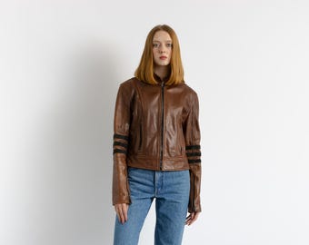 Può includere: Una giacca di pelle marrone con strisce nere sulle maniche. La giacca ha una chiusura con cerniera e un colletto. La modella indossa jeans blu.