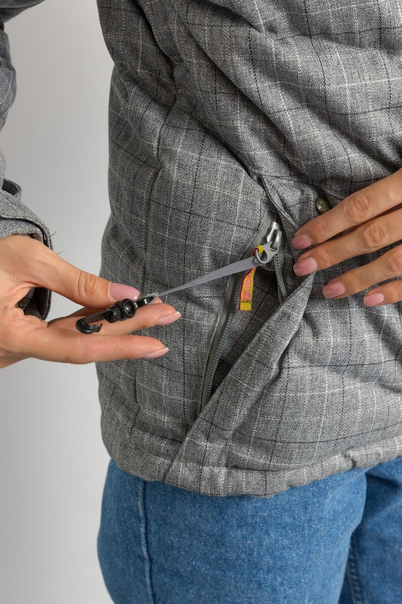 Puede incluir: Primer plano de una chaqueta gris acolchada con un patr&oacute;n de cuadros. La chaqueta tiene cierre de cremallera y un tirador negro. El tirador de la cremallera tiene una etiqueta amarilla y rosa. La persona lleva vaqueros azules.