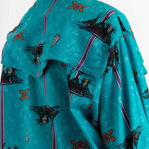 May include: A turquoise blue button-up shirt with a Native American inspired print. The shirt features a repeating pattern of stylized birds, geometric shapes, and stripes in shades of purple, orange, and black.