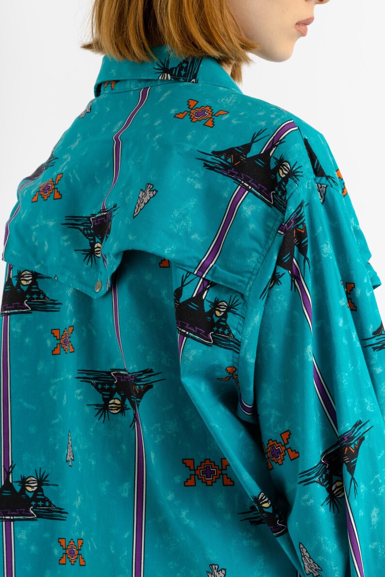May include: A turquoise blue button-up shirt with a Native American inspired print. The shirt features a repeating pattern of stylized birds, geometric shapes, and stripes in shades of purple, orange, and black.