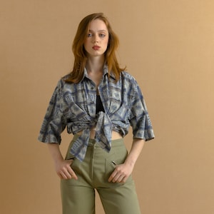 May include: A woman wearing a blue and white patterned shirt tied at the waist and olive green pants.
