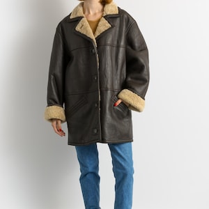 May include: A dark brown leather coat with a beige shearling lining on the collar and cuffs. The coat has a button-up front and two pockets. The person is wearing blue jeans and maroon boots.