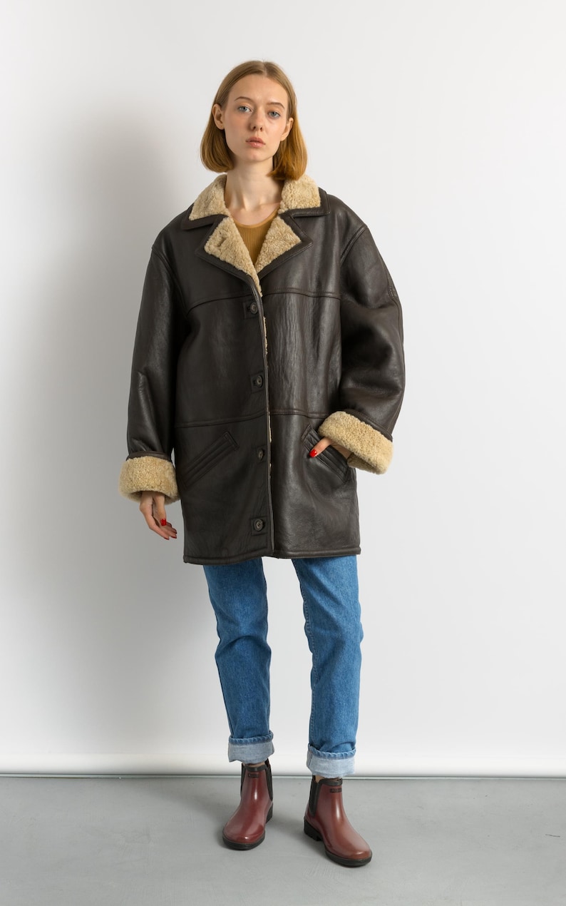 May include: A dark brown leather coat with a beige shearling lining on the collar and cuffs. The coat has a button-up front and two pockets. The person is wearing blue jeans and maroon boots.