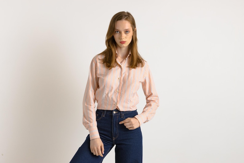 Puede incluir: Una mujer con una camisa de botones a rayas rosa y blancas de manga larga y pantalones vaqueros azules.