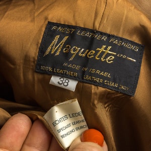 Może przedstawiać: Zbliżenie etykiety odzieżowej marki sk&oacute;rzanej "Moquette". Etykieta jest czarna ze złotymi literami, z napisem "Finest Leather Fashions" i "100% Leather, Made in Israel". Mniejsza etykieta głosi "Echtes Leder". Rozmiar to 38.