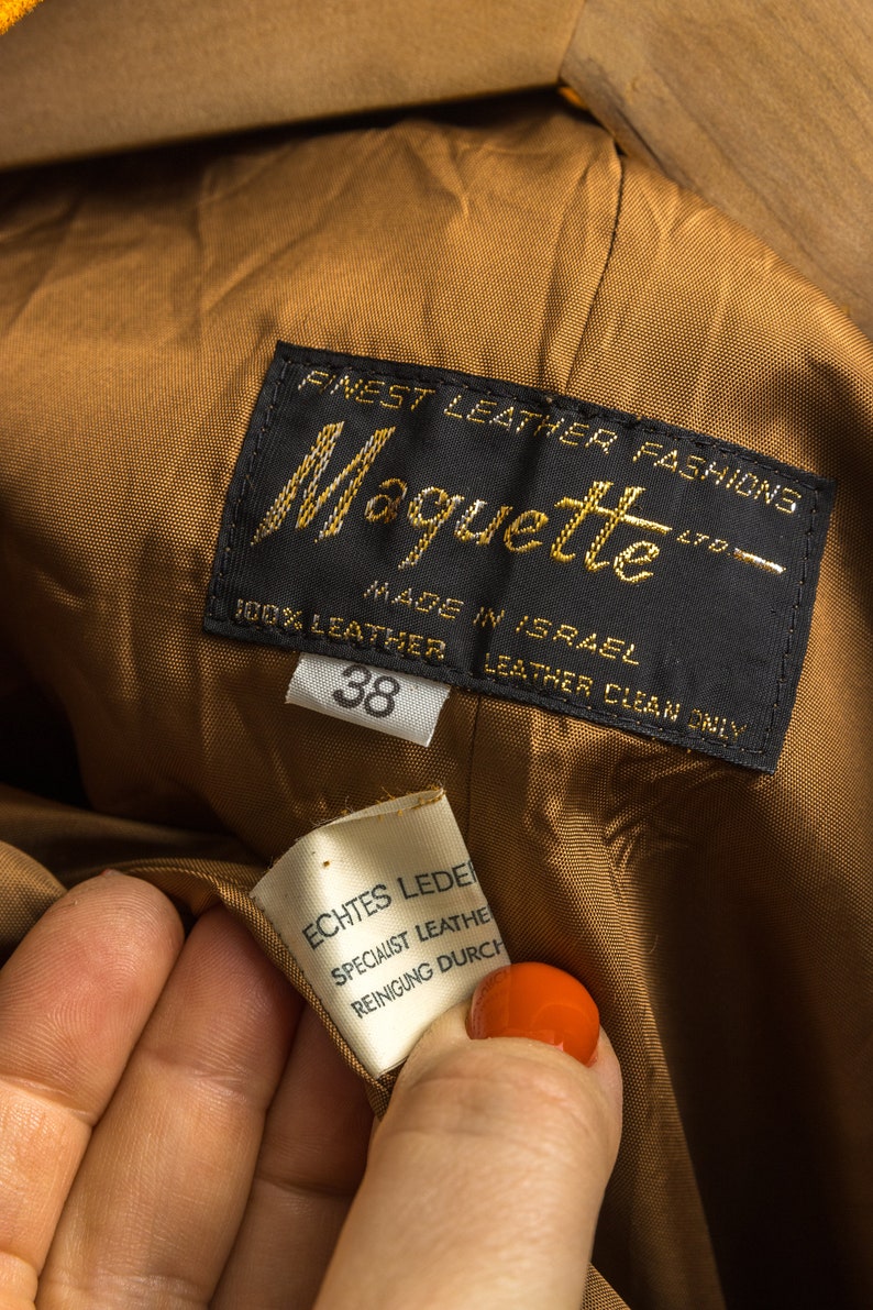 Może przedstawiać: Zbliżenie etykiety odzieżowej marki sk&oacute;rzanej "Moquette". Etykieta jest czarna ze złotymi literami, z napisem "Finest Leather Fashions" i "100% Leather, Made in Israel". Mniejsza etykieta głosi "Echtes Leder". Rozmiar to 38.