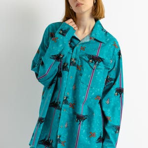 May include: A turquoise blue long-sleeved shirt with a colorful abstract print. The shirt has a button-down collar and a pocket on the left chest.