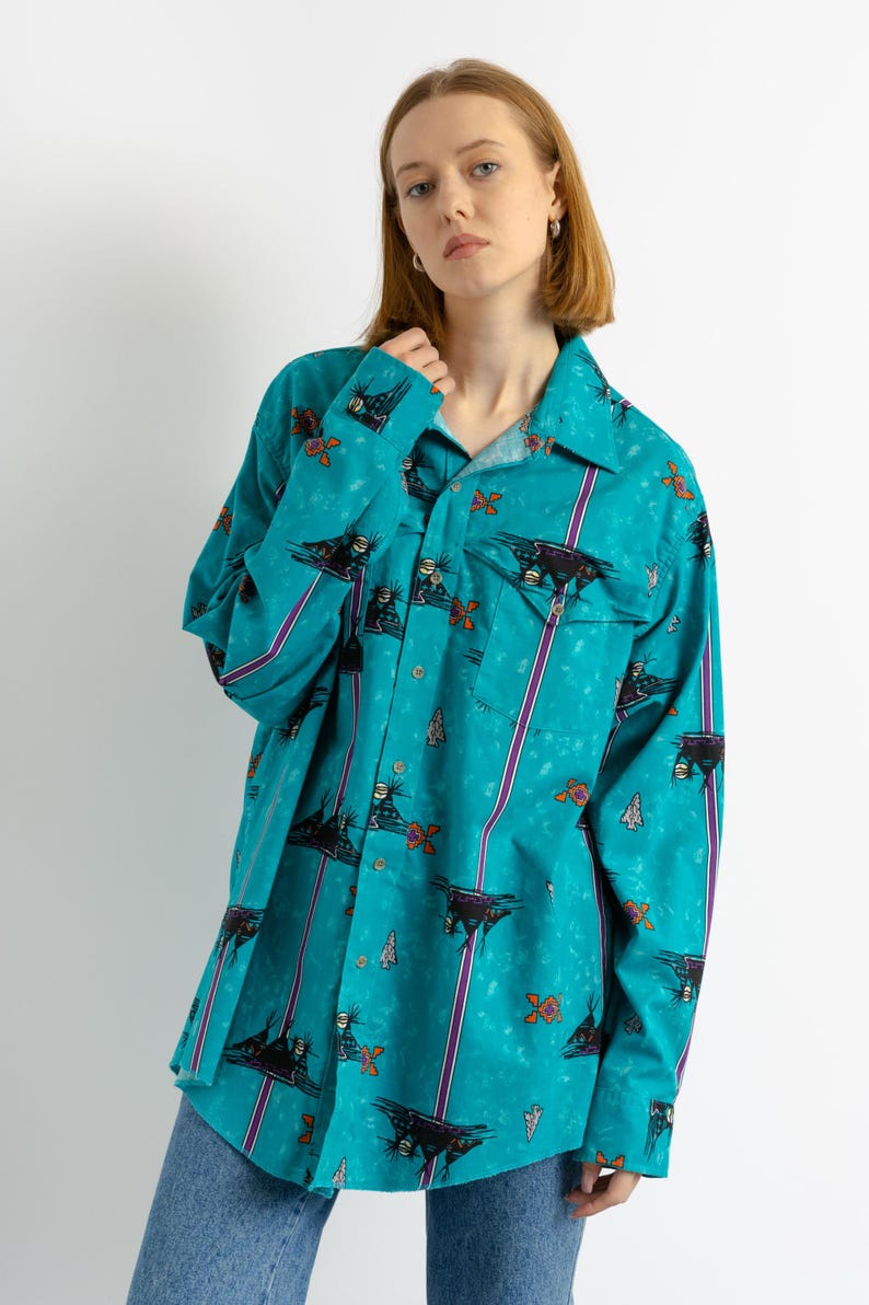 May include: A turquoise blue long-sleeved shirt with a colorful abstract print. The shirt has a button-down collar and a pocket on the left chest.